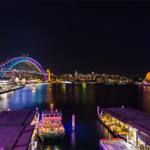 Circular Quay during Vivid Sydney shows the Harbour Bridge in purple and the Opera House in yellow with the north Sydney skyline in the distance.