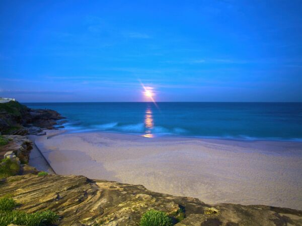 Moonlit reflection on Bronte Beach with rocky cliffs, green vegetation, and golden sand, creating a serene evening atmosphere.