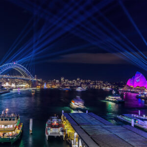 Vivid Sydney photograph showcasing vibrant laser light projections illuminating Circular Quay, stretching from the Harbour Bridge to the Bennelong Apartments. The iconic Opera House glows in pink, set against a dynamic backdrop as ferries glide across the water. Capturing Sydney's landmarks in bold, surreal light, this image celebrates creativity, color, and the city's breathtaking transformation during the annual Vivid festival.