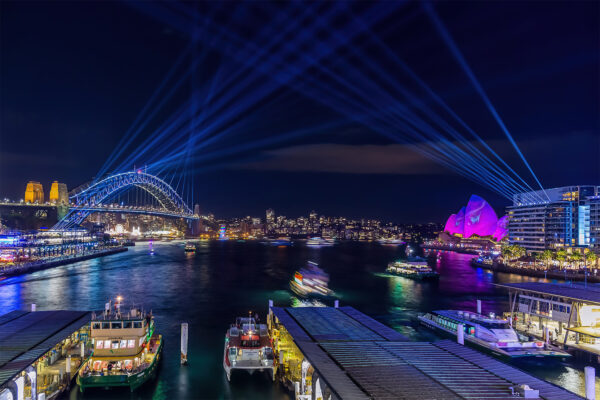 Vivid Sydney photograph showcasing vibrant laser light projections illuminating Circular Quay, stretching from the Harbour Bridge to the Bennelong Apartments. The iconic Opera House glows in pink, set against a dynamic backdrop as ferries glide across the water. Capturing Sydney's landmarks in bold, surreal light, this image celebrates creativity, color, and the city's breathtaking transformation during the annual Vivid festival.