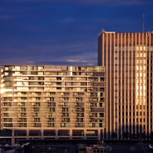 Late afternoon sunlight bathes Bennelong Apartments, also known as the Toaster Building, in a golden glow along Circular Quay in Sydney. Viewed from the third-floor balcony of the Museum of Contemporary Art, the building’s facade is illuminated with warm, rich hues, bringing out depth and vibrant color. A memorable capture of iconic Sydney architecture in dramatic, radiant light.