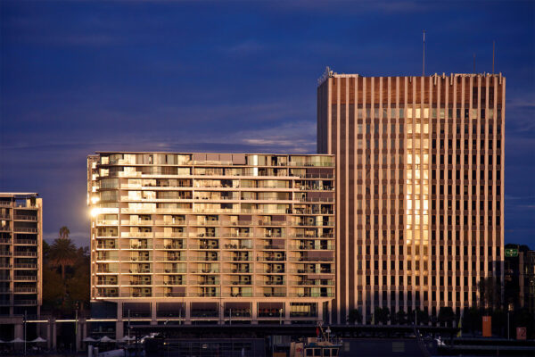 Late afternoon sunlight bathes Bennelong Apartments, also known as the Toaster Building, in a golden glow along Circular Quay in Sydney. Viewed from the third-floor balcony of the Museum of Contemporary Art, the building’s facade is illuminated with warm, rich hues, bringing out depth and vibrant color. A memorable capture of iconic Sydney architecture in dramatic, radiant light.