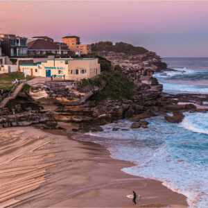 Fine art photograph of Tamarama Beach in Sydney at sunset, featuring soft pink light, gentle waves, freshly groomed sand, and a lone surfer walking from the water.