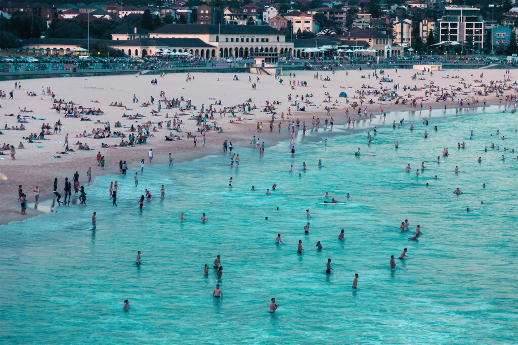 Bondi Beach on a busy summer day, viewed from the north end with the Bondi Pavilion in the background and beachgoers enjoying the ocean.