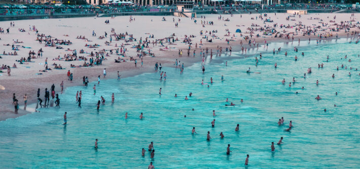 Bondi Beach on a busy summer day, viewed from the north end with the Bondi Pavilion in the background and beachgoers enjoying the ocean.