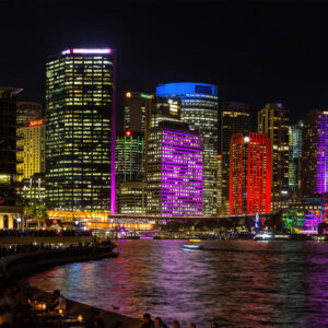 A fine art photograph of Sydney's CBD skyline during Vivid Sydney, taken from Opera Quays. Skyscrapers glow in vibrant purple and red lights, with vivid reflections shimmering across the harbour at night. The image captures the city’s illuminated architecture from a unique perspective, blending light, water, and urban energy.