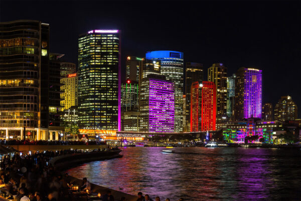 A fine art photograph of Sydney's CBD skyline during Vivid Sydney, taken from Opera Quays. Skyscrapers glow in vibrant purple and red lights, with vivid reflections shimmering across the harbour at night. The image captures the city’s illuminated architecture from a unique perspective, blending light, water, and urban energy.