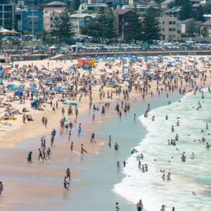 A summer day at Bondi Beach with people in the ocean, sunbathing on the sand, and relaxing under blue umbrellas, with coastal apartments and pine trees in the background.