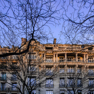 Classic Haussmann-style Paris apartment building with ornate balconies and tall windows, framed by bare winter tree branches and lit by warm afternoon sunlight in the Trocadéro district.