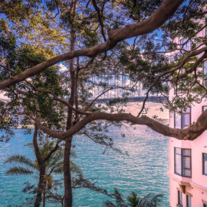 Sydney Harbour viewed through overhanging tree branches in Kirribilli, with turquoise harbour water, a pink Art Deco apartment building and the Sydney Harbour Bridge visible in the background.