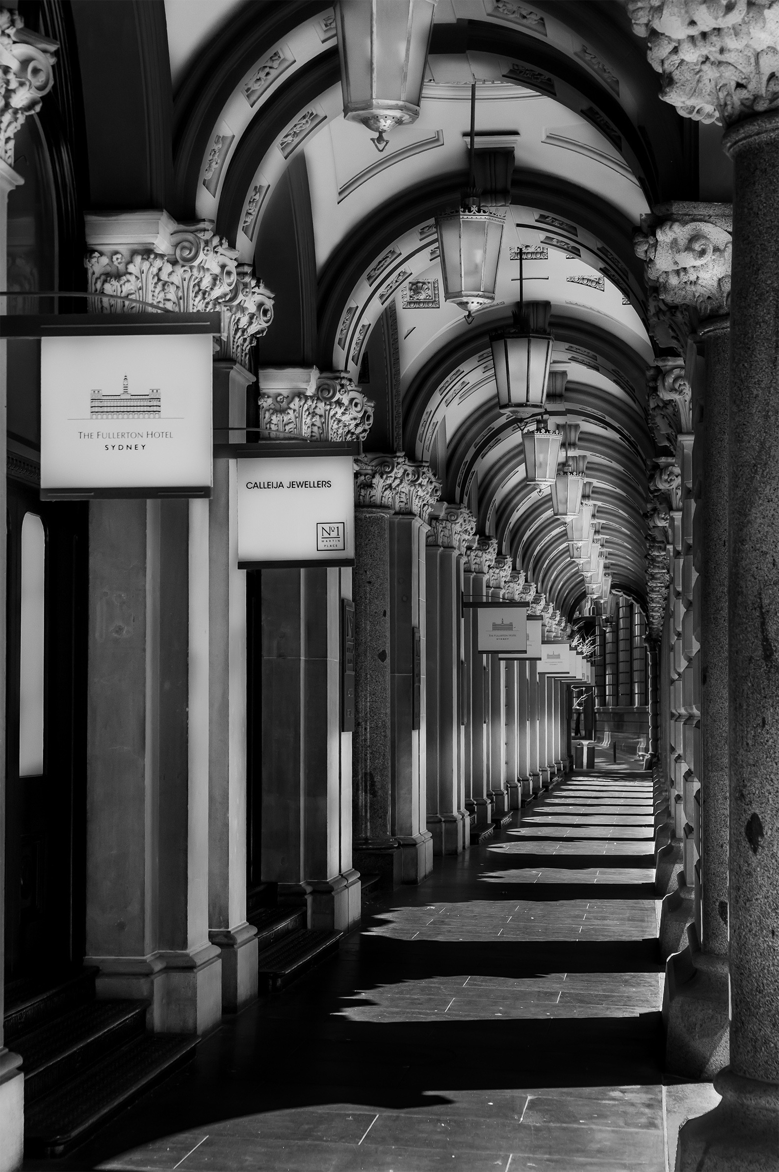 Black and white architectural photograph of The Colonnades in Martin Place, Sydney, featuring ornate sandstone columns, repeating arches, hanging lanterns and dramatic sunlight casting geometric shadows along the historic walkway.